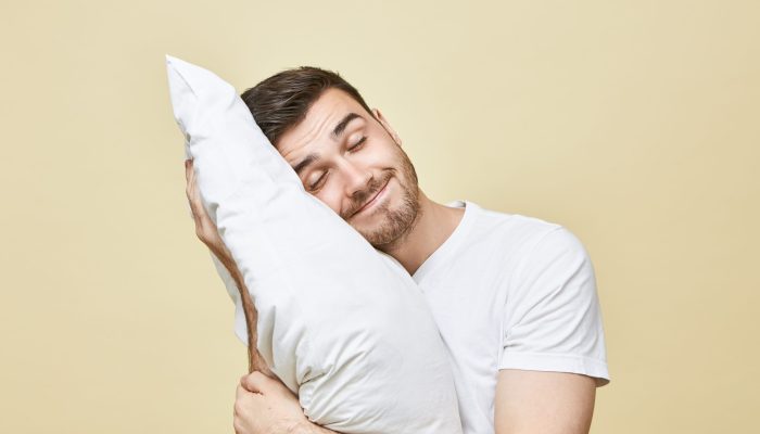 Horizontal image of handsome cute young man with bristle posing with head on white soft pillow sleeping peacefully and smiling, seeling good dream. Attractive guy napping after hard working day