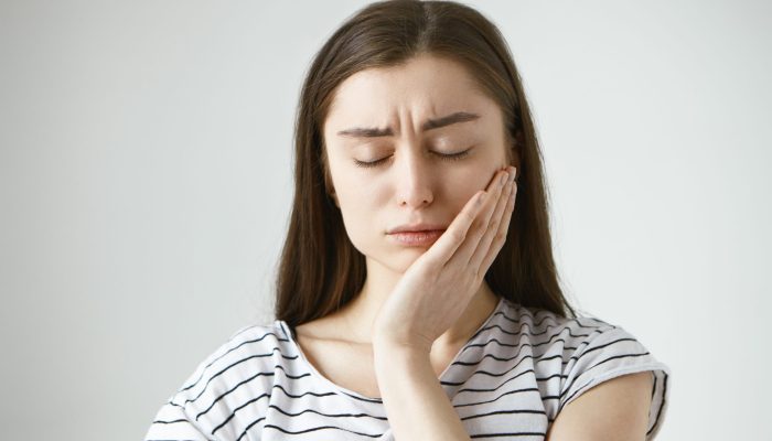 Portrait of unhappy depressed young Caucasian brunette woman closing eyes in frustration and pressing hand against cheek, trying to soothe pain while suffering from toothache, having tooth decay